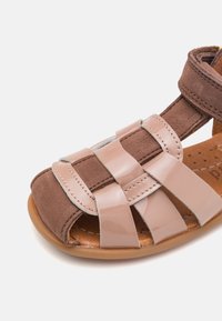 Brązowe zamszowe sandały z błyszczącymi różowymi paskami. Posiadają otwarty przód i tył, z przeszyciami oraz jasnobrązową gumową podeszwą.