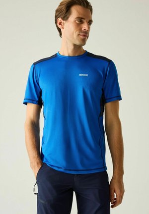 Mężczyzna w niebieskiej sportowej koszulce z krótkim rękawem i ciemnych spodniach, stojący z jedną ręką w kieszeni, patrzący w bok na jednolitym tle.