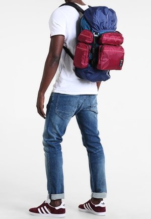 Mand iført en blå og rød rygsæk med flere lommer, hvid t-shirt, blå jeans og røde sneakers med hvide striber, står sideværts.