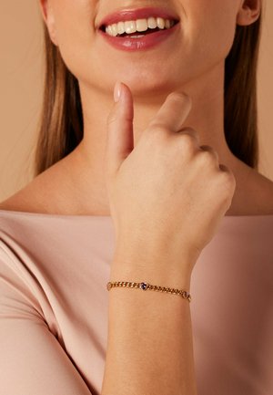 Glimlach met zichtbare tanden en lippen met lichtroze lippenstift, waarbij een pols wordt getoond die versierd is met een gouden armband met kleine paarse hartvormige bedels.