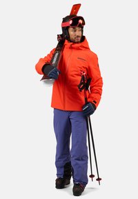 Heldere oranje skijas met zwarte accenten, paarse skibroek, zwarte skischoenen, houdt skies en stokken vast, draagt een helm en skibril.