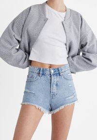 Mujer vestida con shorts de mezclilla claros desgastados, top corto blanco y chaqueta gris de botones, con las manos en las caderas frente a un fondo blanco.