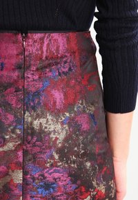 Gros plan d'une personne portant un pull côtelé foncé et une jupe taille haute arborant un motif floral abstrait éclatant dans des tons de rouge, rose, bleu et or.
