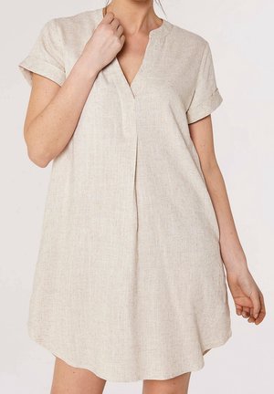 Femme portant une robe en lin beige clair, à manches courtes, ample, avec un petit col en V et des manches retroussées, debout devant un fond uni.