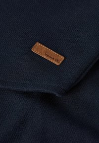 Tessuto a maglia di un blu navy scuro con un'etichetta rettangolare in pelle marrone cucita che recita "name it" sulla superficie piegata.