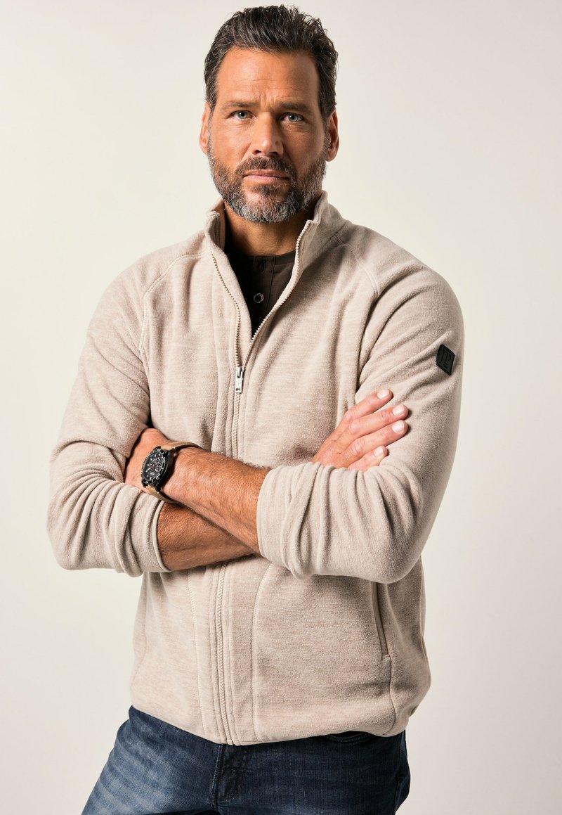 Chaqueta de forro polar cremosa con cierre de cremallera y cuello alto, fabricada con tejido texturizado y un acento de logo negro en el hombro izquierdo. Llevada sobre una camiseta oscura.