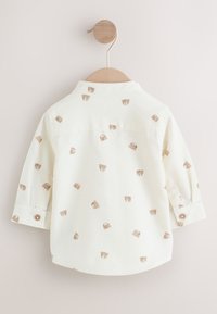 Chemise en coton blanc avec un ourlet arrondi, ornée de dessins de tête d'ours marron clair, manches longues et poignets à boutons. Suspendue sur un cintre en bois.