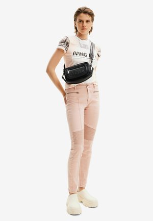 Schwarze Umhängetasche mit geprägtem Logo, rosa schmale Jeans mit gerippten Akzenten und beigen klobigen Plateau-Schuhen. Person steht selbstbewusst.