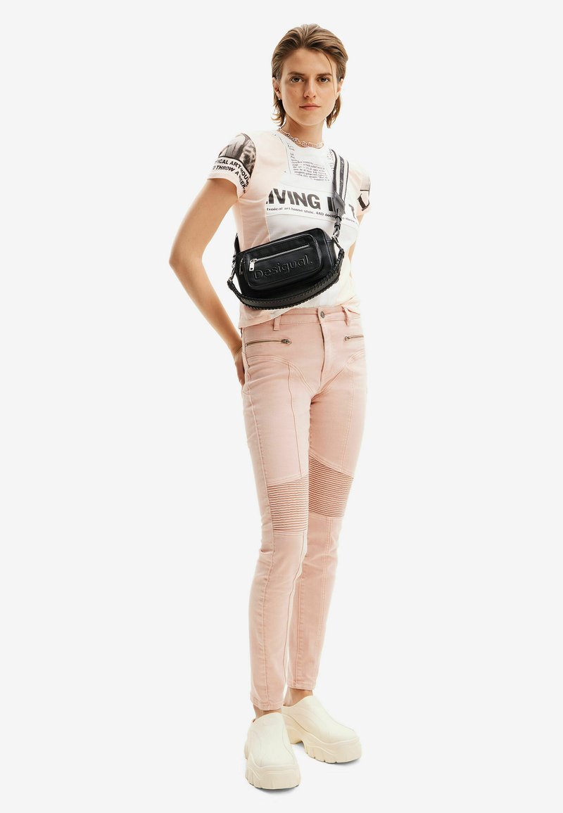 Svart axelremsväska med präglad logotyp, rosa snäva jeans med ribbade detaljer och beige klumpiga plattformsskor. Personen står självsäkert.