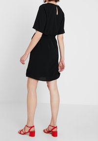 Zwarte jurk met korte mouwen, lichte glans, boven de knie lengte, met een sleutelgat aan de achterkant, gecombineerd met rode hoge hakken.