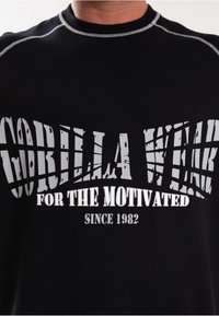 Zwarte T-shirt met korte mouwen, voorzien van een gedurfde witte tekst die luidt: "GORILLA WEAR FOR THE MOTIVATED SINCE 1982," met een geribbelde textuur.