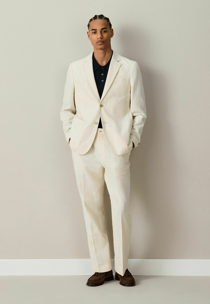 Homme aux cheveux tressés portant un costume couleur crème, une chemise noire et des chaussures marron, debout contre un mur uni avec les mains dans les poches.