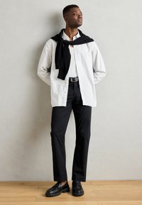 Cămașă albă cu dungi, butonată, peste un tricou negru, combinată cu pantaloni negri și mocasini negri. O geacă neagră aruncată pe gât.