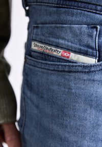 Pantalones de mezclilla azules con tirantes que presentan un bolsillo frontal con una etiqueta visible de "Diesel Industry". Tejido texturizado y diseño clásico con herrajes de metal.