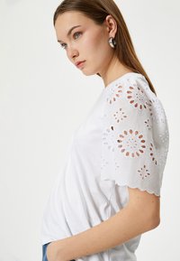 Hvid bomulds t-shirt med broderede korte ærmer, der har blomstermønstre med udskæringer og bølgede kanter. Enkel og afslappet pasform.