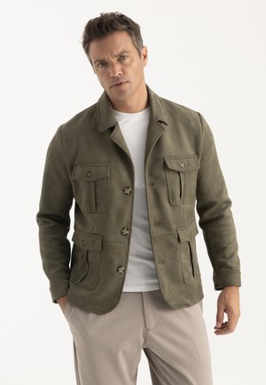 Homme portant une veste boutonnée vert olive sur une chemise blanche et un pantalon beige, debout devant un fond uni.