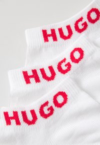 Bílé bavlněné ponožky s červeným logem "HUGO", žebrovaná textura v oblasti lemu, naskládány ve třech vrstvách s výraznými kontrastními barvami.