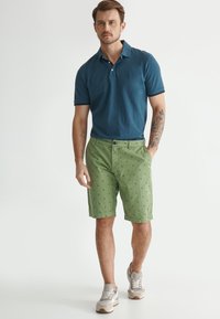 Polo shirt teal à manches courtes avec des coutures blanches, associé à un short vert présentant un petit motif marine. Des baskets grises complètent le look.