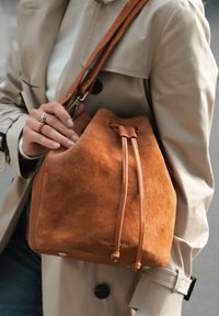 Braune Wildleder-Eimerhandtasche mit glatten Lederakzenten, verstellbarem Tragegurt und Kordelzugverschluss. Sichtbare Markenkennzeichnung an der Unterseite.