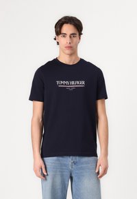Marineblå bomulds t-shirt med korte ærmer og rund hals. Har "TOMMY HILFIGER" logo i hvidt med røde og blå accenter centreret på brystet.