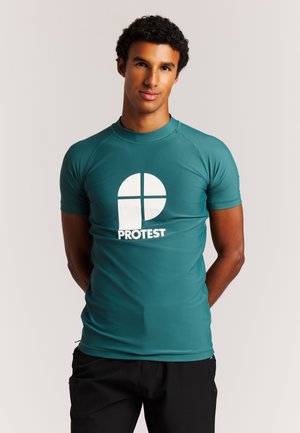 Jonge man staat met handen achter de rug, draagt een getailleerd teal shirt met een groot wit "PROTEST"-logo op de voorkant.