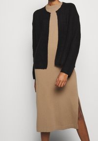 Cardigan nero a maglia sopra un vestito beige, con collo tondo e spacco laterale. Il cardigan ha una texture morbida e una vestibilità ampia.