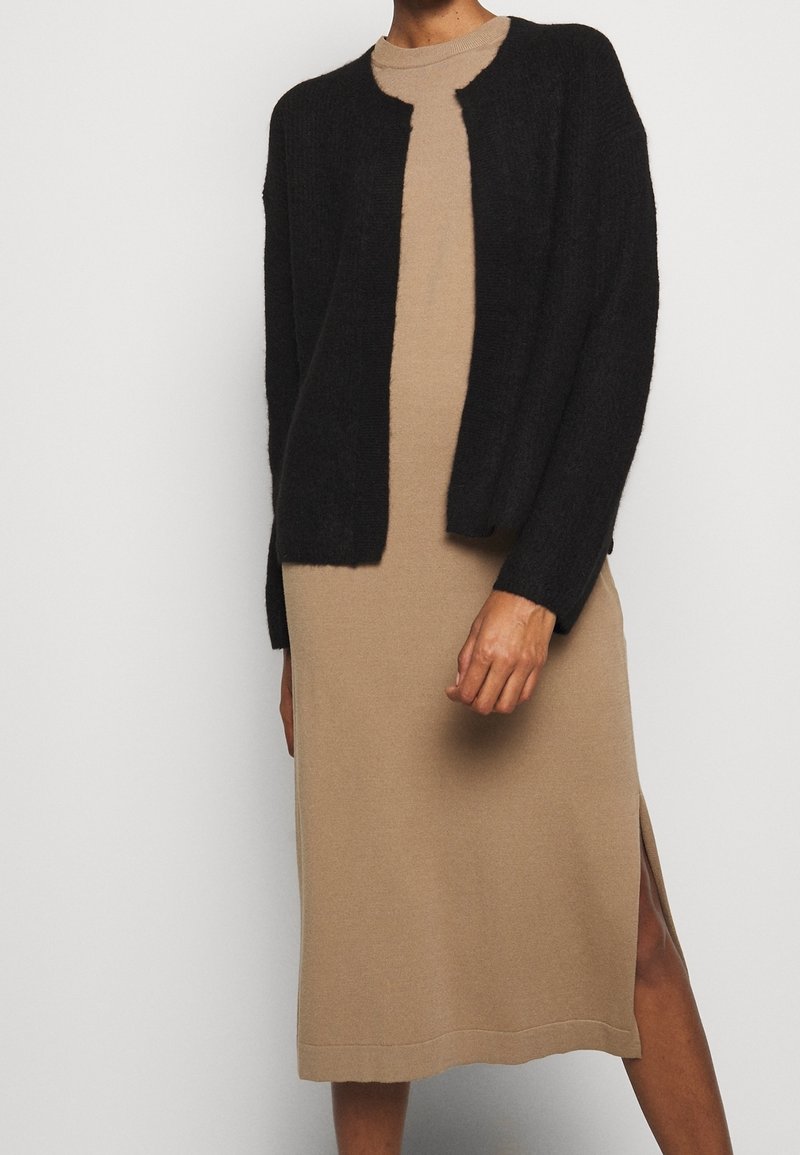 Cardigan nero a maglia sopra un vestito beige, con collo tondo e spacco laterale. Il cardigan ha una texture morbida e una vestibilità ampia.