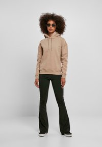 Beige hettegenser med snorer, løs passform, sammen med mørke vide bukser og svarte sneakers. Modell med solbriller foran en ensfarget bakgrunn.