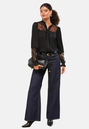 Schwarze Bluse mit Spitzenakzenten, langen Ärmeln und einer Knopfleiste, kombiniert mit weit geschnittenen, dunkelblauen Jeans und spitzen schwarzen Stiefeln.