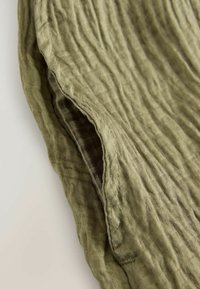 Gros plan d'un tissu froissé vert olive avec des coutures visibles et une texture suggérant un matériau doux et léger.