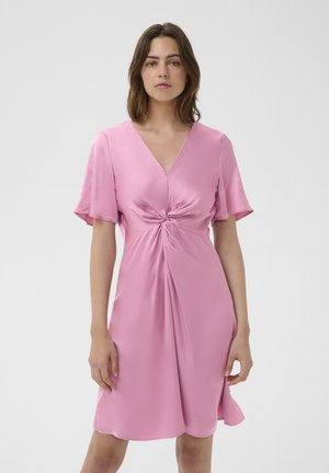 Nő, aki térdig érő rózsaszín szatén ruhát visel, rövid legyezős ujjakkal és csavart csomó részlettel a deréknál, egy egyszínű háttér előtt áll.