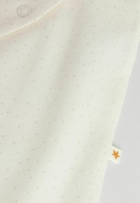 Baumwollstoff in Creme mit hellbraunen Pünktchen, ausgestattet mit einem runden Kragen und einem kleinen orangefarbenen Sternetikett an der Seite.