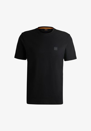 T-shirt preta feita de algodão suave. Apresenta um decote redondo e mangas curtas. Um pequeno logótipo texturizado em forma de quadrado está posicionado no lado esquerdo do peito.