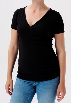 Kobieta ubrana w dopasowaną czarną koszulkę z krótkim rękawem i dekoltem w serek oraz jasnoniebieskie dżinsy, stojąca na tle białego, jednolitego tła.