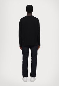 Camisetta nera a maniche lunghe, vestibilità ampia, con scollo rotondo; abbinata a jeans scuri e sneakers bianche, vista da dietro.