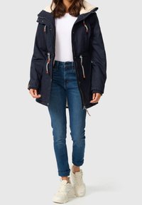 Granatowa parka z kapturem wyścielanym sztucznym futrem, ozdobiona troczkami, zamkami błyskawicznymi i brązowymi akcentami, noszona na białej bluzce i niebieskich dżinsach.