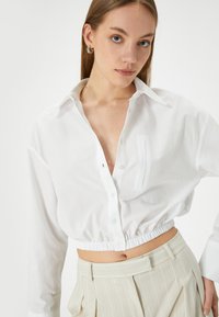 Camicia bianca a maniche corte con colletto, con tasca frontale e orlo elastico. Indossata con pantaloni plissettati di colore chiaro.