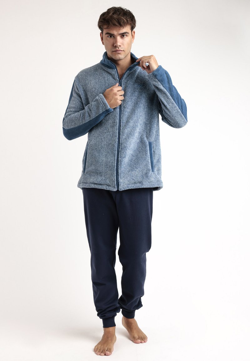 Chaqueta de forro polar azul con mangas en un azul oscuro que contrastan, dos bolsillos laterales, cuello alto y cremallera completa. Combinada con pantalones de chándal azul marino.