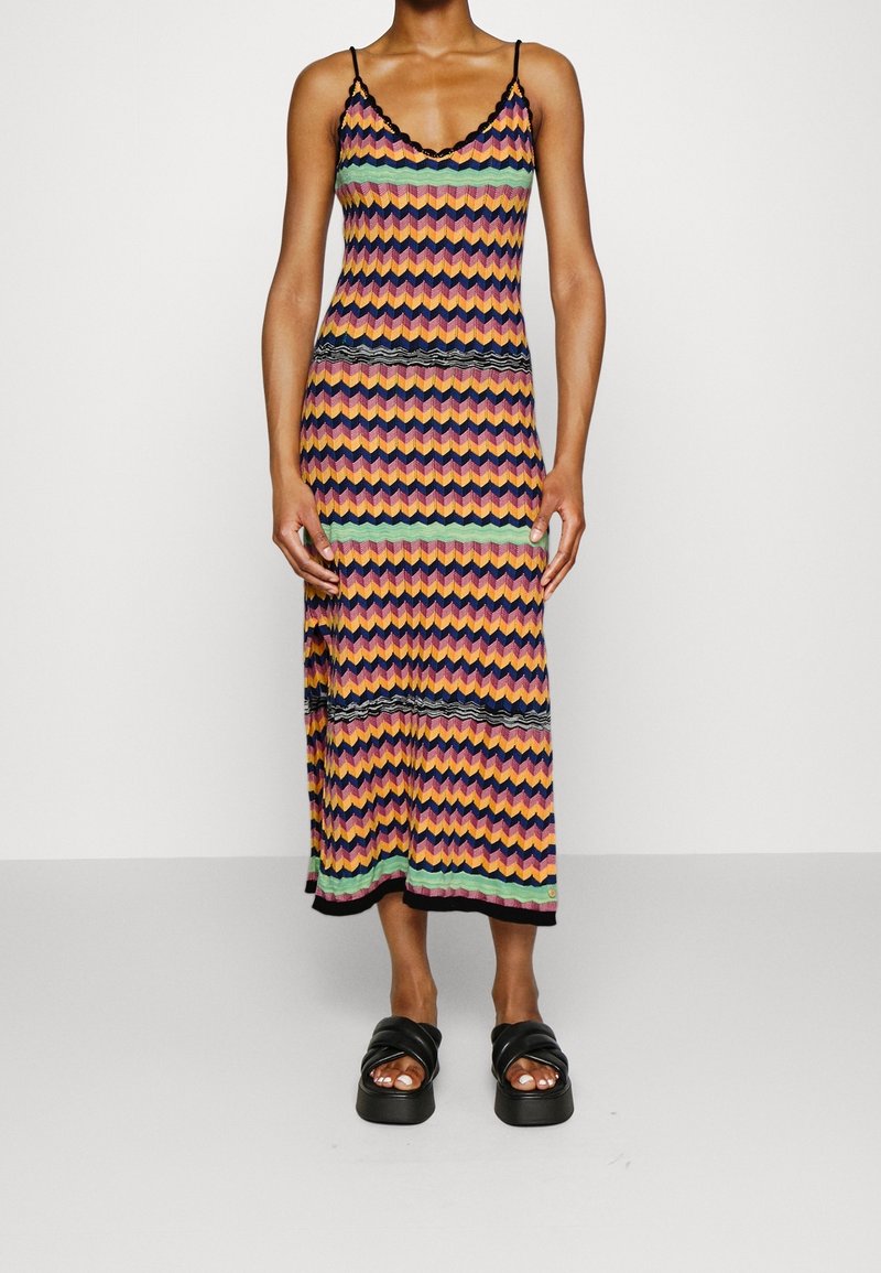 Femme portant une robe mi-longue sans manches à motif zigzag multicolore et des sandales à plateforme noires, debout devant un fond uni.