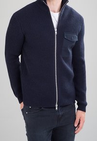 Granatowy sweter na zamek z dzianiny o prążkowanej fakturze, z wysokim kołnierzem i kieszenią na lewej piersi. Posiada srebrny zamek błyskawiczny oraz dopasowane mankiety.