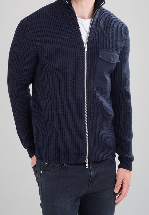 Granatowy sweter na zamek z dzianiny o prążkowanej fakturze, z wysokim kołnierzem i kieszenią na lewej piersi. Posiada srebrny zamek błyskawiczny oraz dopasowane mankiety.