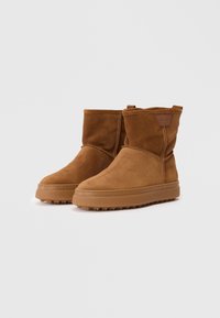 Ruske nubuk ankle botte med rund tå, bløt tekstur og gummisåle med gripeplugger. Inkluderer en merket etikett på siden.