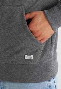 Sudadera gris con bolsillo tipo canguro, ribeteado en el dobladillo y los puños, y una etiqueta blanca en la parte inferior izquierda delantera que indica la marca y las instrucciones de cuidado.