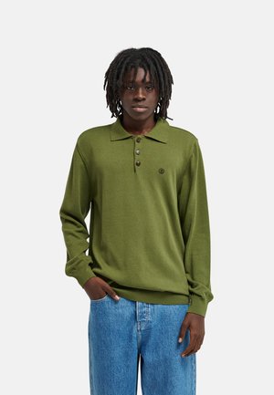 Giovane con dreadlocks fino alle spalle che indossa un maglione polo verde oliva a maniche lunghe e jeans blu, in piedi davanti a uno sfondo semplice.
