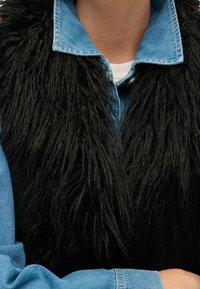 Svart fuskpälsväst lager på lager över en denimskjorta. Mjuk, ludden textur kontrasterar med den strukturerade denim. Blå och svart färgkombination är framträdande.