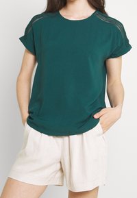 T-shirt à manches courtes bleu sarcelle avec un col rond et un bord en dentelle transparente sur les épaules, associée à un short plissé beige clair.