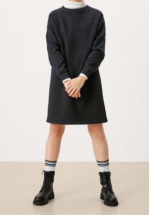 Robe sweatshirt noire à coupe décontractée, avec des poignets côtelés et un col rond, associée à des chaussettes blanches montantes avec des rayures noires et des bottes de combat.