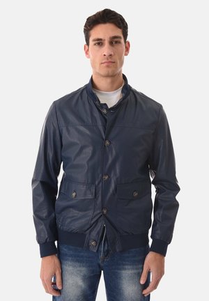 Giovane uomo che indossa una giacca blu navy con bottoni, tasche frontali e polsini a costine, abbinata a jeans blu sbiaditi su uno sfondo bianco semplice.