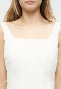 Γυναίκα που φοράει λευκό αμάνικο φόρεμα με υφασμένα λουλουδένια σχέδια και τετράγωνο ντεκολτέ, εμφανιζόμενη από τους ώμους μέχρι το πάνω μέρος του στήθους.