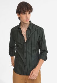 Camisa de botones a rayas verde oscuro, con cuello, mangas largas y corte ajustado. Presenta una tapeta frontal y un sutil brillo en la tela.
